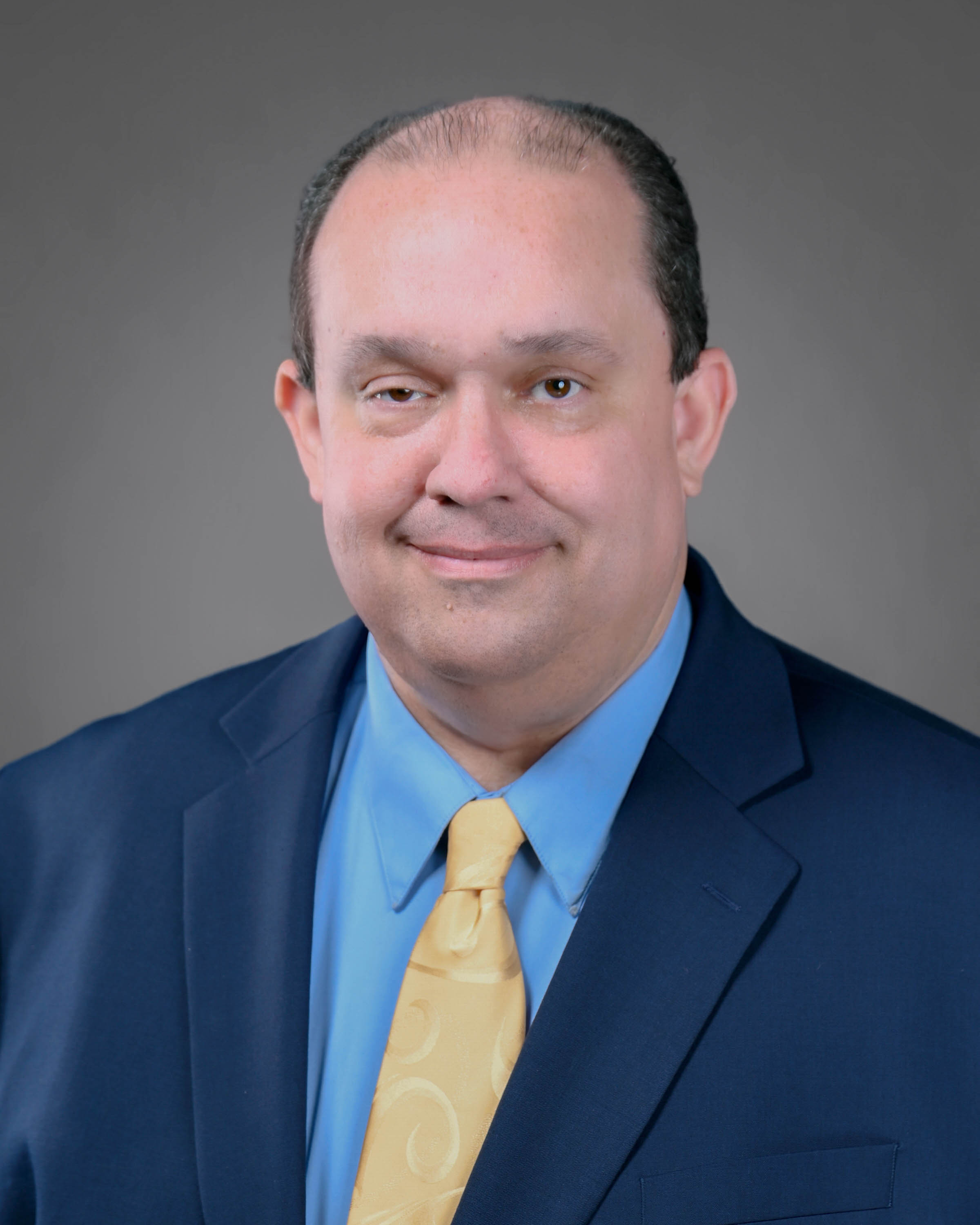 Smiling man that is clean shaven wearing a blue suit with a light blue shirt and yellow power tie.